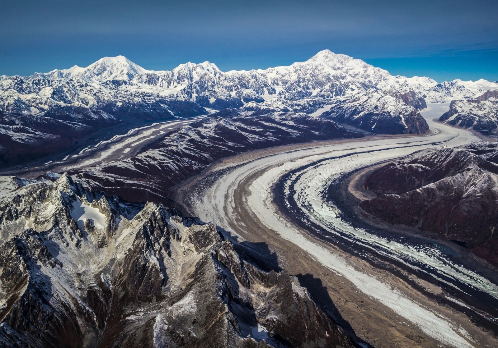 Wide aerial view of branching glaciers curling through dark ridges beneath a panorama of snow-covered mountains.
