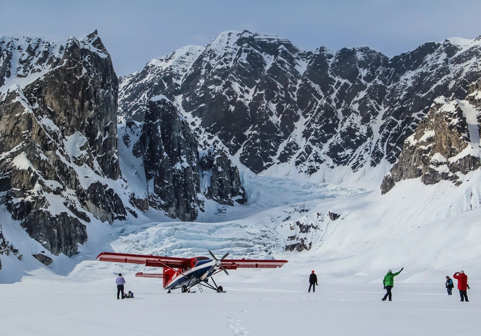 A red-wing ski airplane and scattered visitors on a broad snowy glacier below towering dark mountain walls and a blue icefall.