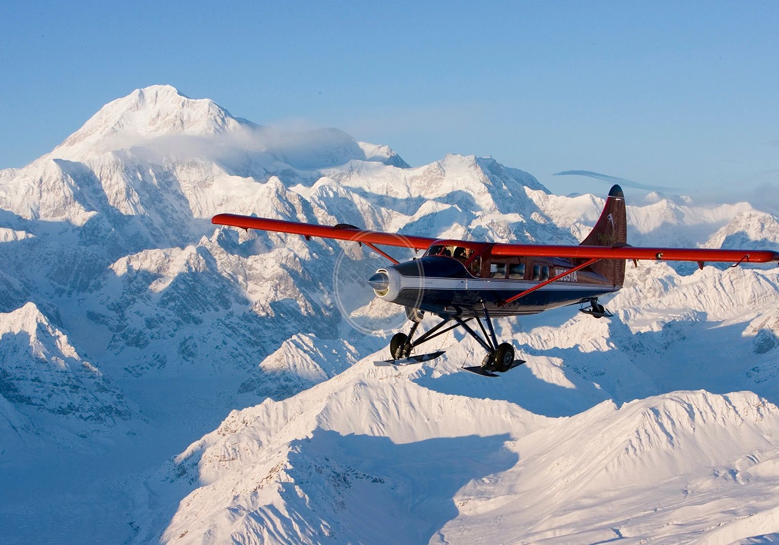 Side-on red-wing, blue-and-white ski airplane flying in front of bright snow-covered peaks beneath a pale blue sky.