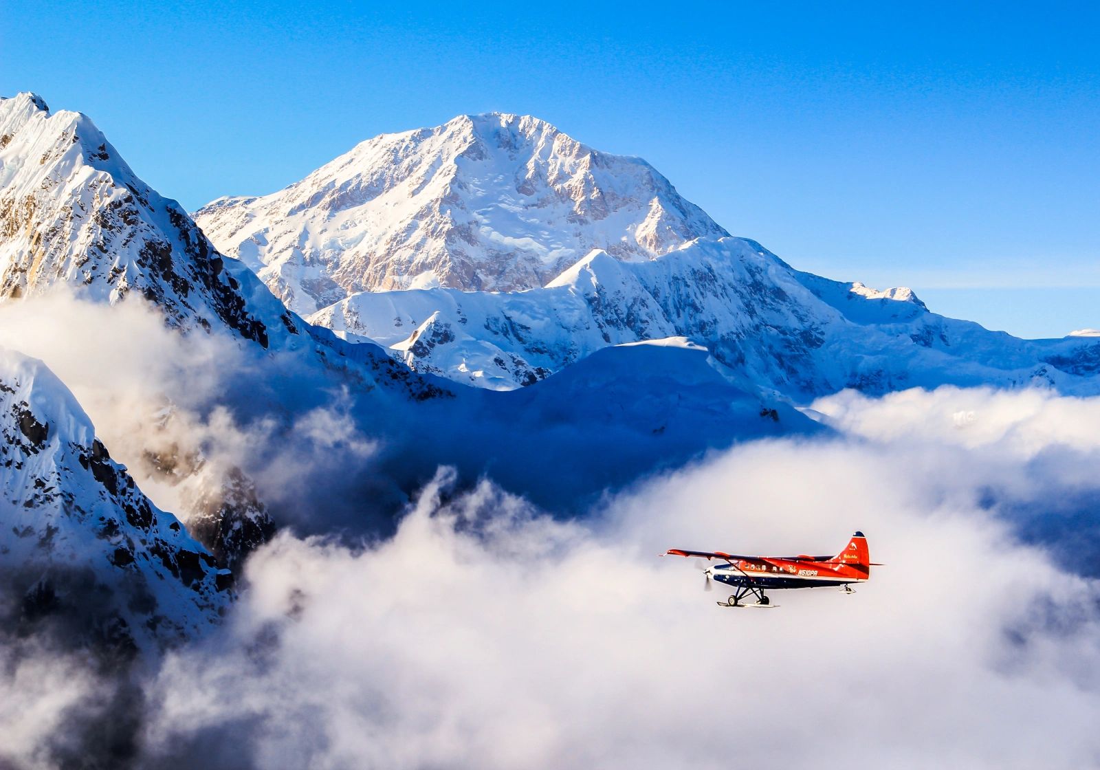 A small red-wing airplane flies at lower right among white clouds beneath a massive sunlit snowy mountain and saturated blue sky.