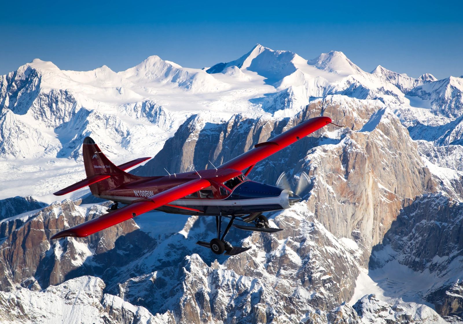 Large red-wing, blue-and-white airplane flying beside sunlit sheer rock walls and snowy peaks under a vivid blue sky.