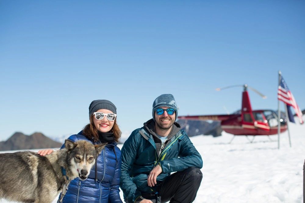 Two smiling visitors pose with a husky in snow, with a red helicopter and an American flag behind them.