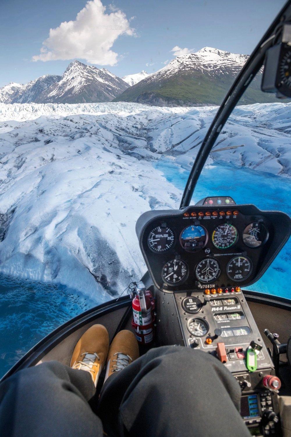 Vertical passenger-eye view through a helicopter cockpit over fractured white glacier ice and turquoise melt pools, with mountains ahead.