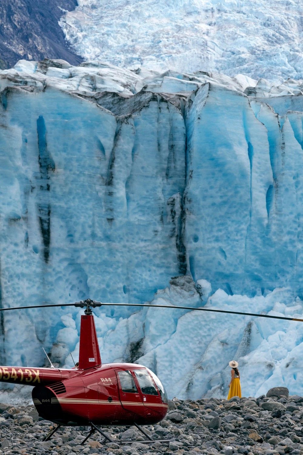 Vertical composition of a red helicopter on rocky ground beneath a tall blue ice wall; a visitor in a yellow dress stands beside the glacier.