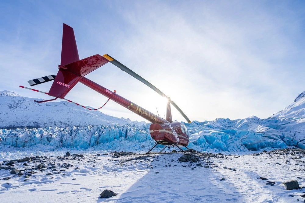 Red helicopter seen from behind, landed near bright blue fractured glacier ice with mountains and low sun behind its rotor.