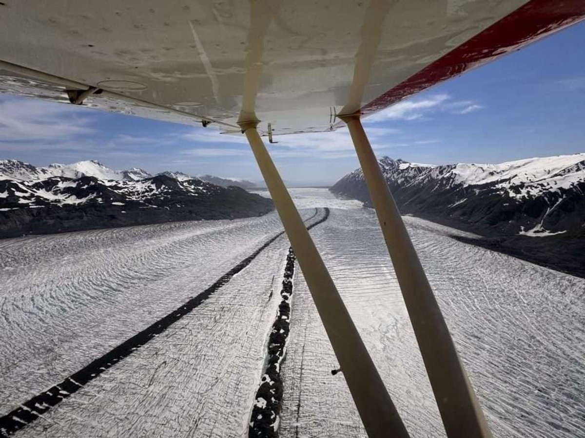 View beneath an airplane wing across a wide glacier with dark moraines between snowy mountains.