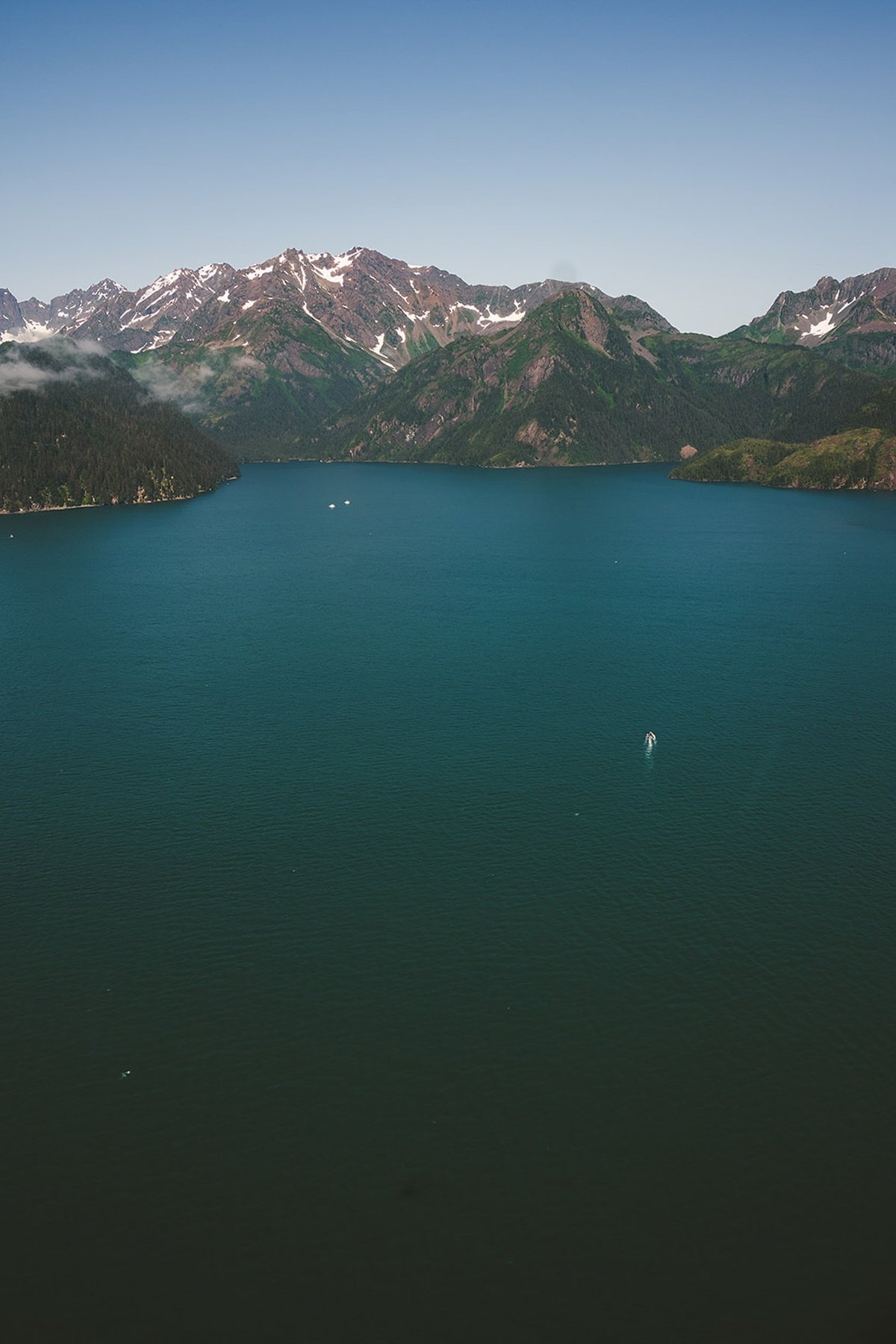 Aerial view across deep-blue water toward forested coastal mountains with patches of snow.