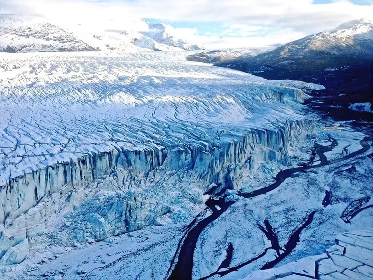 Aerial view of a blue-white glacier front and dark branching channels below snow-covered mountains.